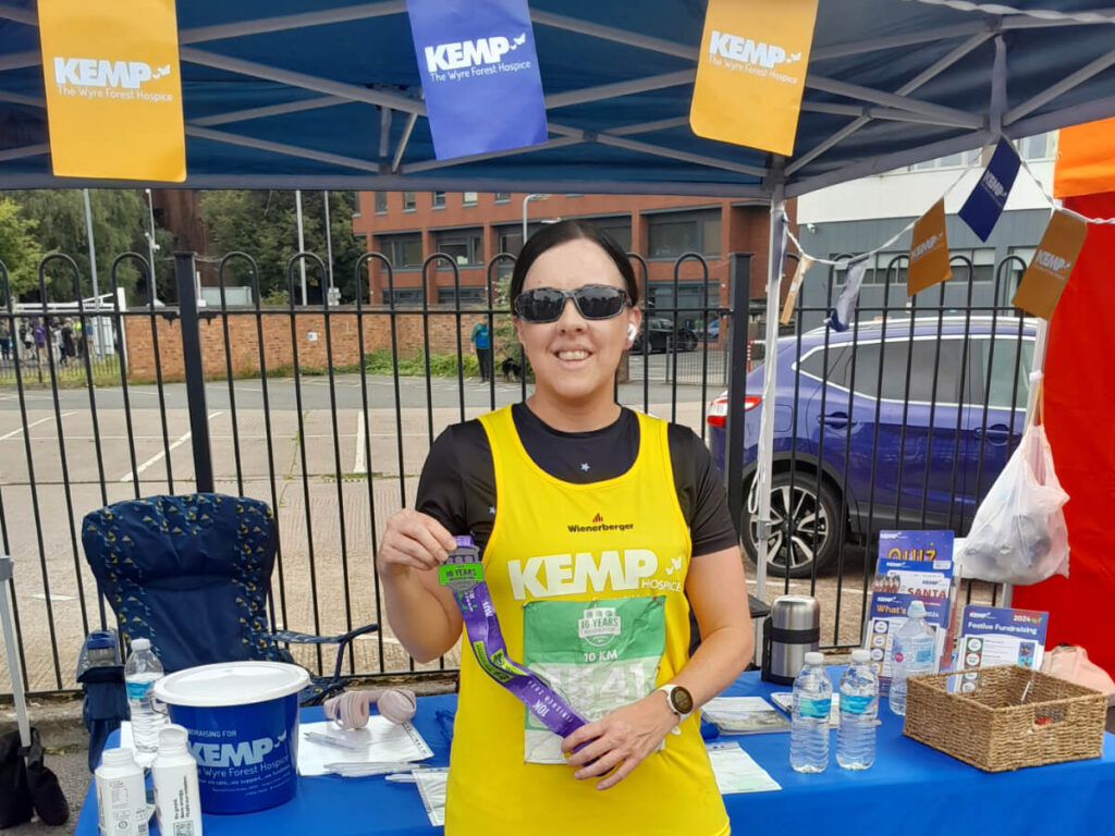 a lady stands wearing a yellow running vest infront of a table with kemp brand materials on