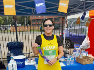 a lady stands wearing a yellow running vest infront of a table with kemp brand materials on