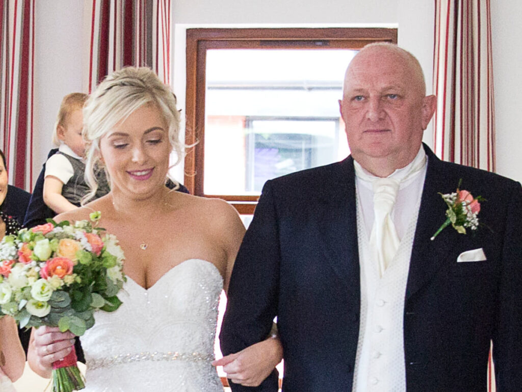 a lady stand in a wedding dress walking down the aisle holding arm in arm with a male in a suit