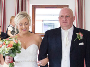a lady stand in a wedding dress walking down the aisle holding arm in arm with a male in a suit