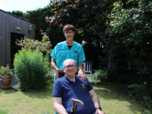 a man sits outside in a garden with a nurse wearing scrubs stood behind