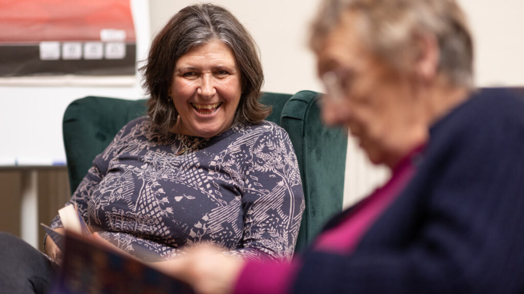 a lady sits smiling by another lady who is blurred in the foreground