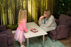 a lady sits in a purple armchair talking to a young girl standing opposite across a table. She wears a pink jumper and skirt