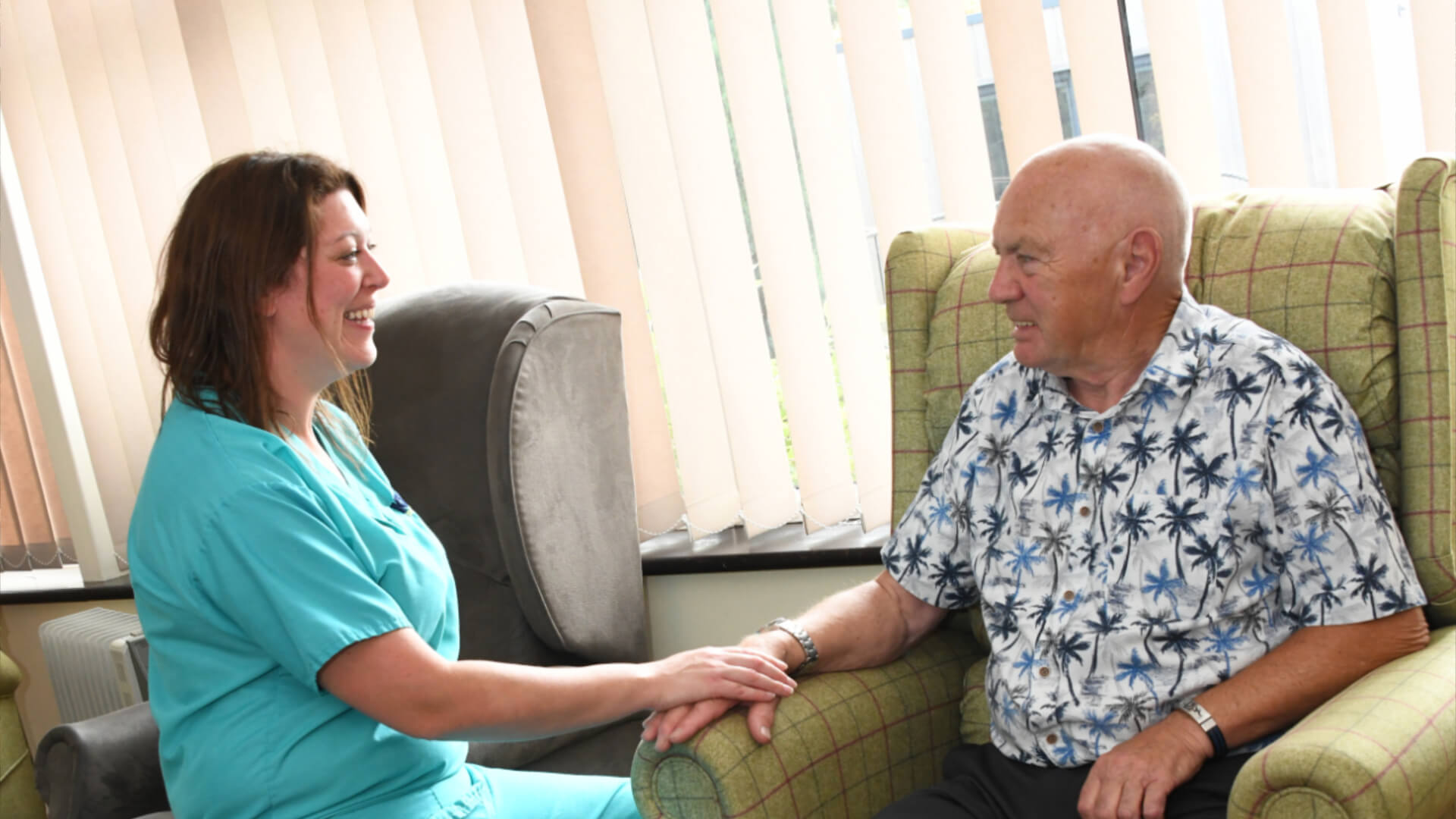 a nurse in scrubs sits opposite a patient hand in hand in a supportive manner