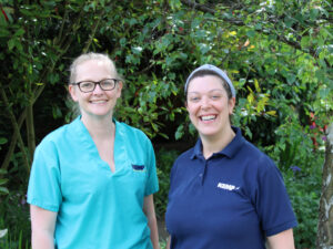 A nurse stands in scrubs and another stands in a blue tshirt outside in front of a green tree