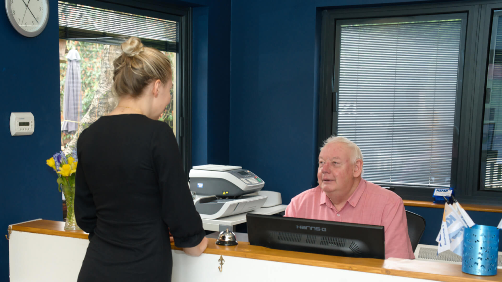 a gentleman sits behind a reception desk talking to a lady