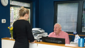 a gentleman sits behind a reception desk talking to a lady