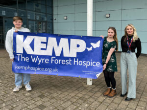 two students stand holding a kemp hospice blue and white banner with another lady stood outside a school building
