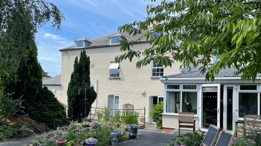 a cream building with a conservatory door open with trees ans shrubbery in the foreground