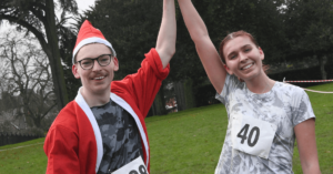 Two people stood with one hand raised, one wears a Santa suit and another with a racing number.