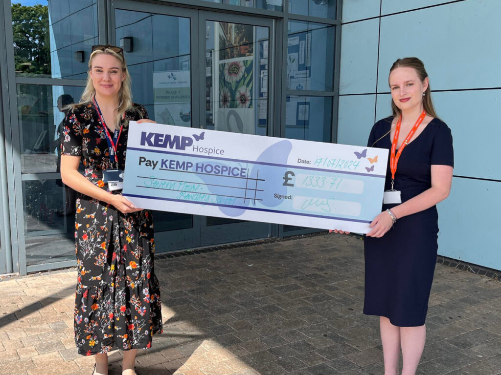 two ladies stand holding a large cheque outside a school