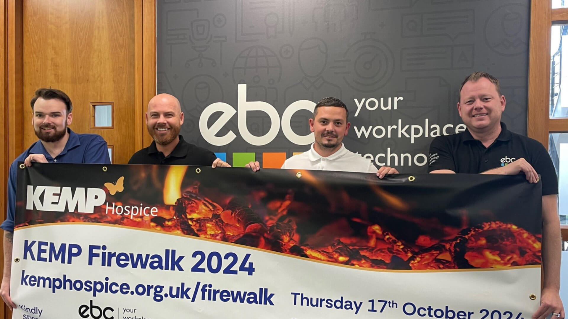 four people stand in an office holding a vinyl banner to promote the kemp firewalk