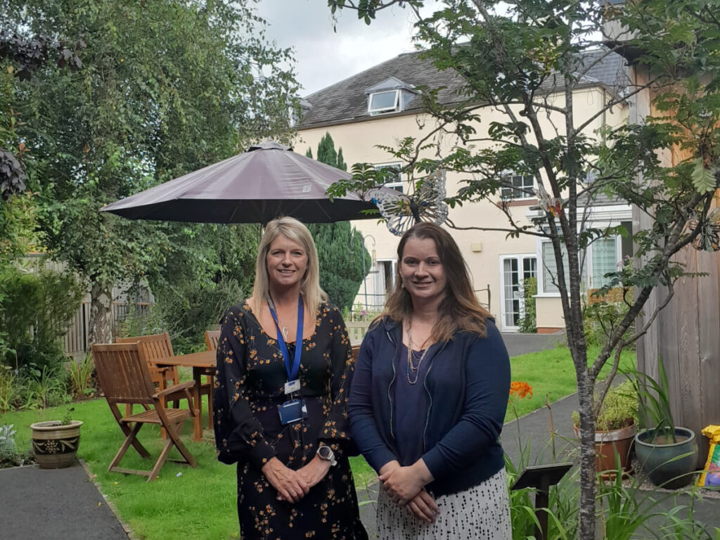 two ladies stand in a garden area next to a tree and with a picnic table and umbrella behind them.