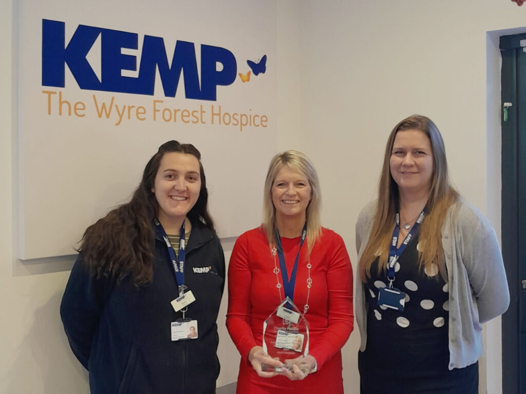 three ladies stand together. The lady in the middles wears a red dress and holds a glass award trophy