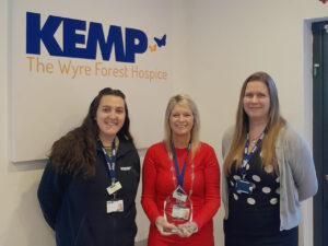 three ladies stand together. The lady in the middles wears a red dress and holds a glass award trophy