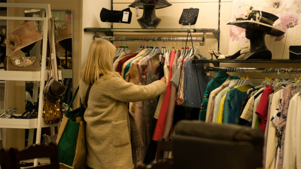 a lady in a white coat stands looking at a rail of clothes in a shop