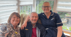 a lady sits to the left, she is smiling with a gentleman sat in the middle with a nurse wearing dark blue scrubs sat to the right. They all look happy.