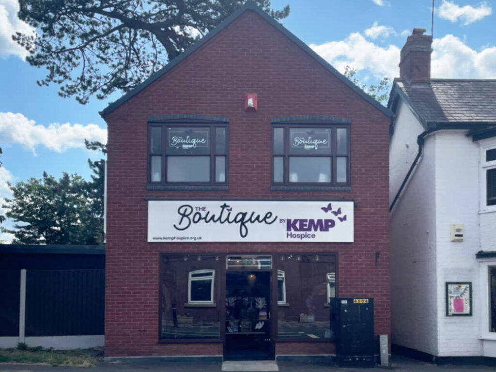 a brick building on a high street showing new bright signage with the words boutique by kemp hospice. The building has dark window frames with two windows upstairs and two downstairs.