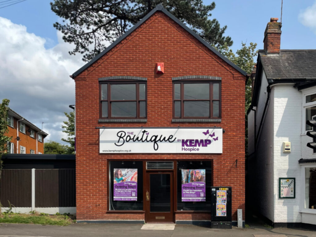a brick building on a high street showing new bright signage with the words boutique by kemp hospice. The building has dark window frames with two windows upstairs and two downstairs.