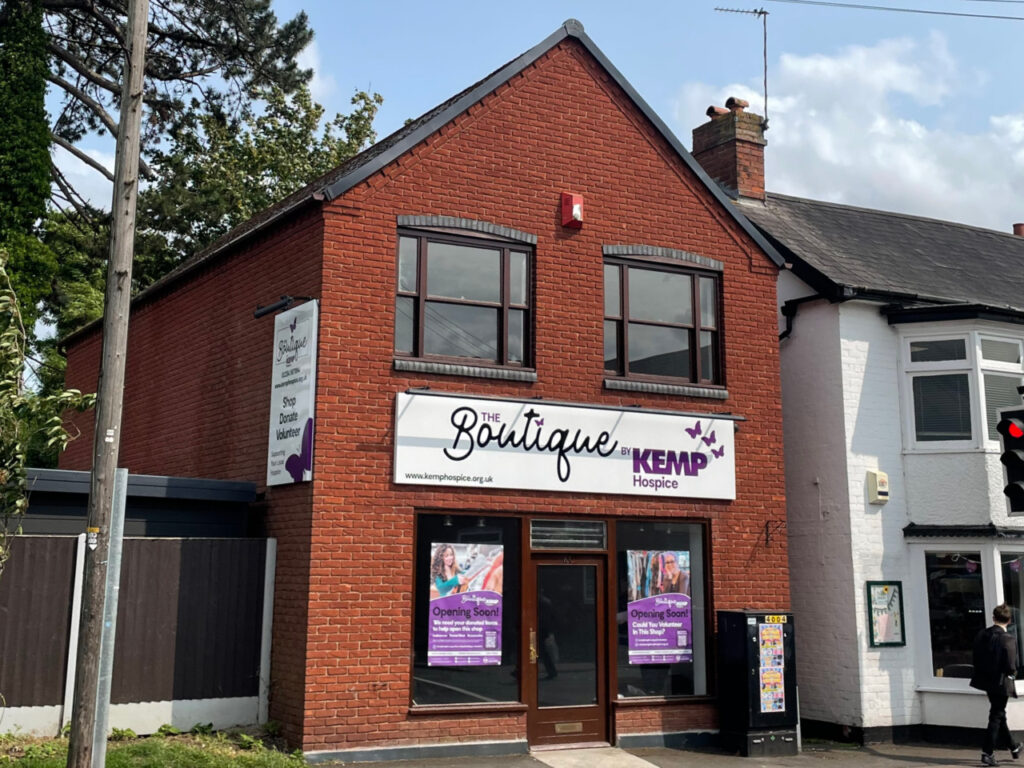 a brick building on a high street showing new bright signage with the words boutique by kemp hospice. The building has dark window frames with two windows upstairs and two downstairs.