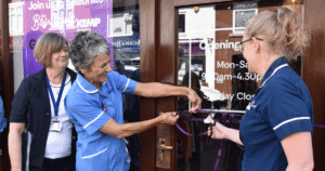 two ladies wearing KEMP nurse tops and another lady stand together and cut a ribbon on a door