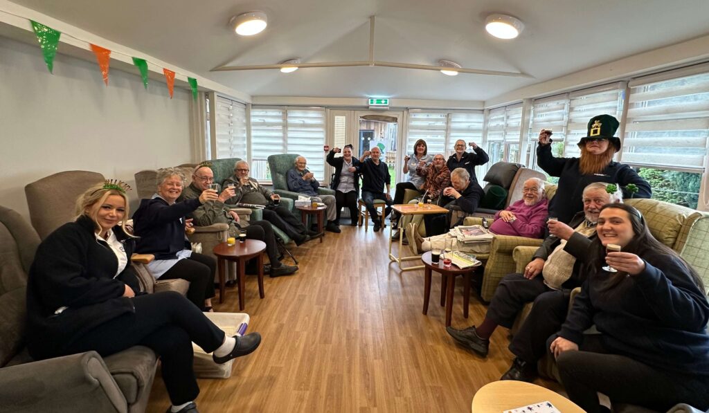 A group of people sitting together smiling with a drink in hand cheering for st Patricks Day