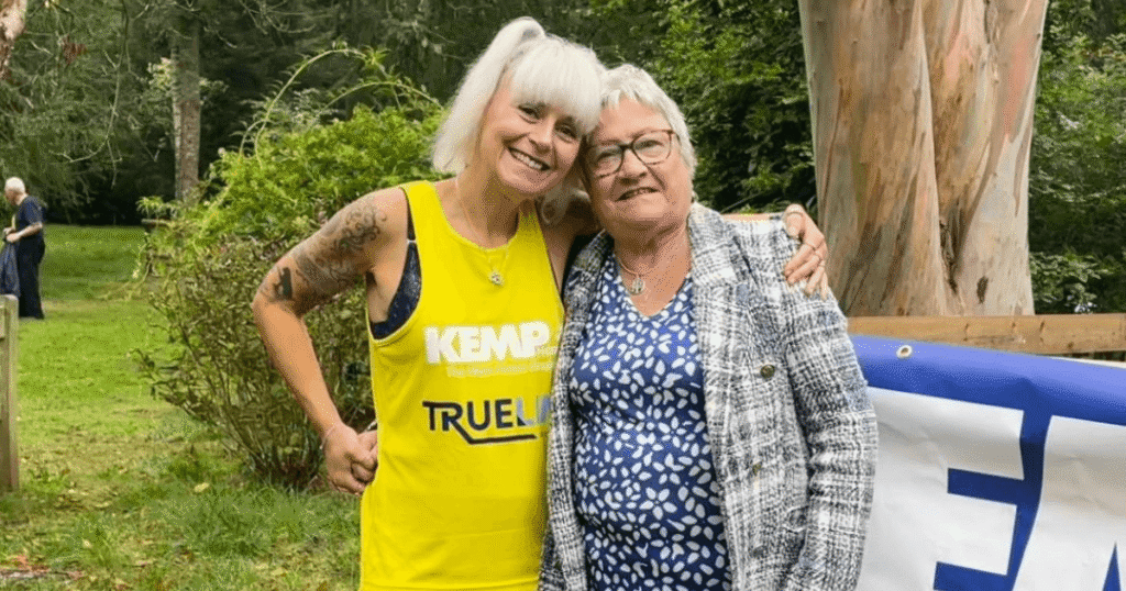 two ladies stand arm in arm. One wears a yellow running vest and the other wears a blue and white dress with a checked jacket. They are standing in a wooded area with a tree and bush in the background.