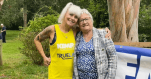 two ladies stand arm in arm. One wears a yellow running vest and the other wears a blue and white dress with a checked jacket. They are standing in a wooded area with a tree and bush in the background.