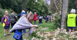 a lady takes a photo of a butterfly sitting on flowers in a bush with other peoples standing in the background.