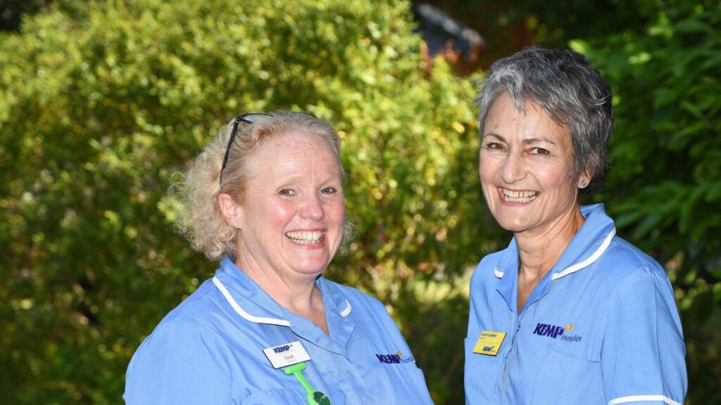 Two nurses stand together in a garden with greenery in the background