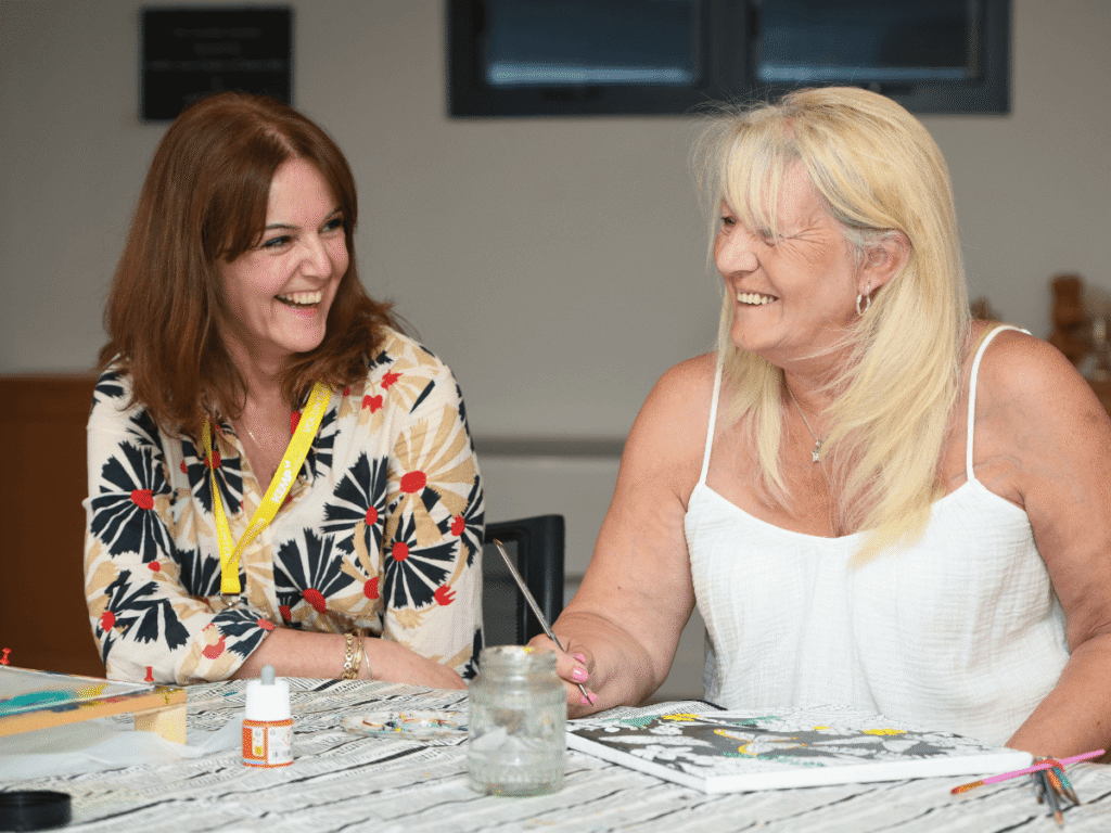 two ladies sit at a table smiling. One is holding a paint brush by a canvas she is painting on.