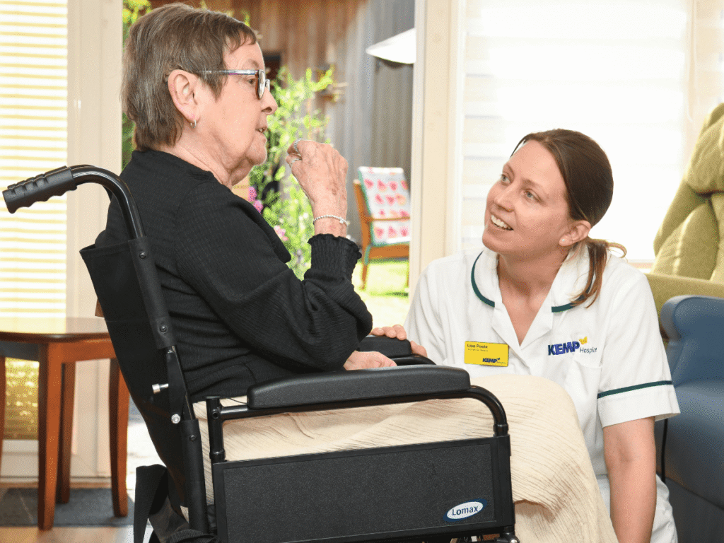 a lady sits in a wheelchair while another lady wearing white scrubs is knelt down talking to her.