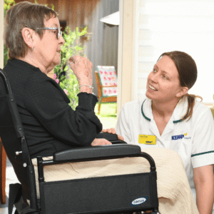 a lady sits in a wheelchair while another lady wearing white scrubs is knelt down talking to her.