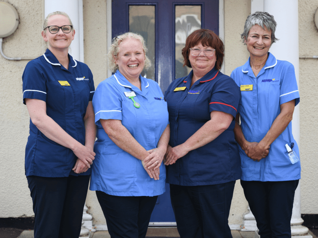 a team of four nurses wearing scrubs stand in a line smiling and looking at the camera.