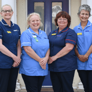 a team of four nurses wearing scrubs stand in a line smiling and looking at the camera.