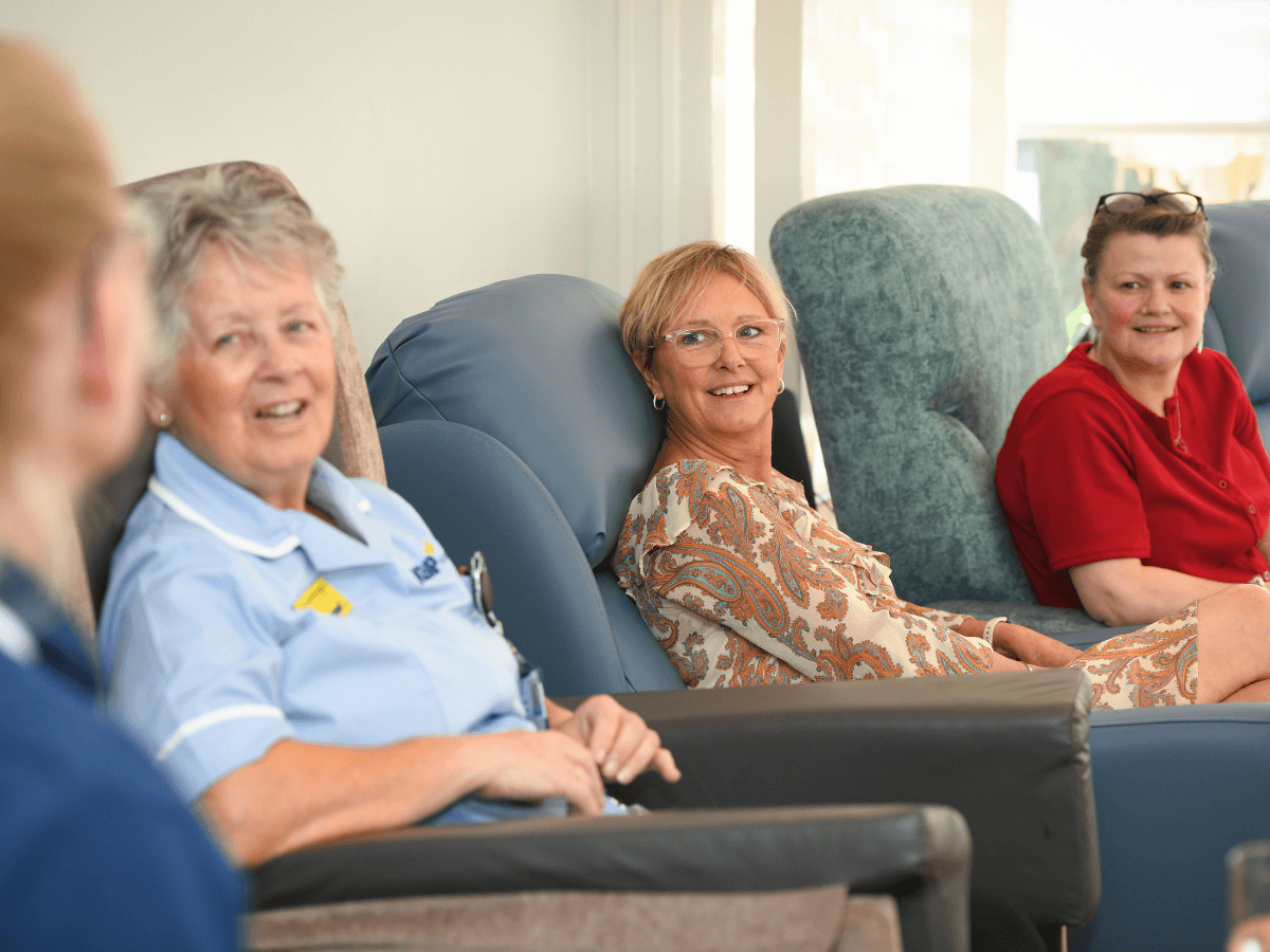 a nurse in light blue scrubs and a lady in an orange and cream dress sit in blue armchairs looking towards another nurse to the side of the camera, both are smiling.