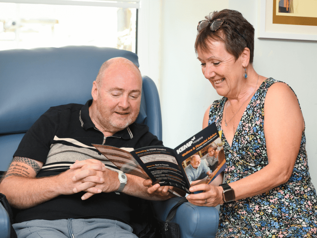a lady sits next to a gentleman sat in a blue chair smiling looking through a leaflet.
