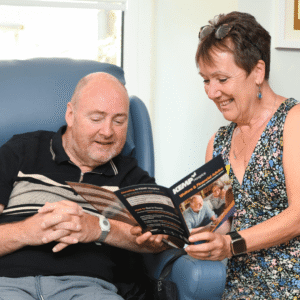 a lady sits next to a gentleman sat in a blue chair smiling looking through a leaflet.