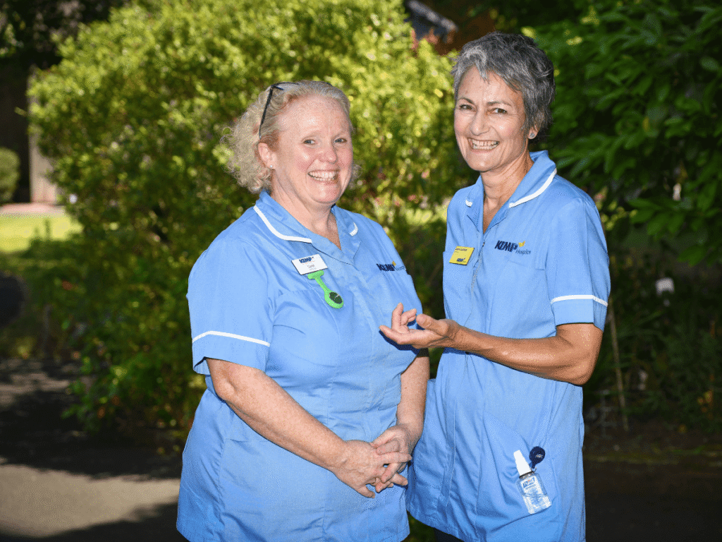 two nurses wearing light blue scrubs stands smiling looking directly at the camera with green shrubbery blurred in the background on a sunny day.