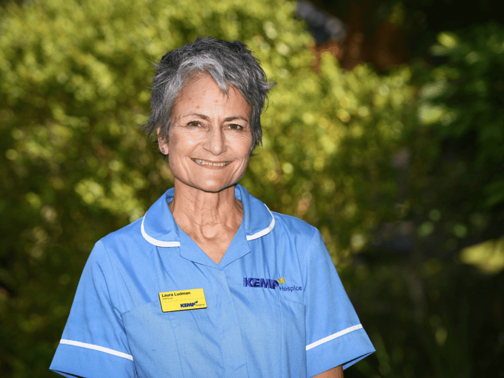 a nurse wearing light blue scrubs stands smiling looking directly at the camera with green shrubbery blurred in the background on a sunny day.
