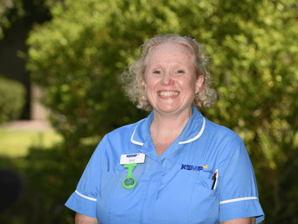a nurse wearing light blue scrubs stands smiling looking directly at the camera with green shrubbery blurred in the background on a sunny day.