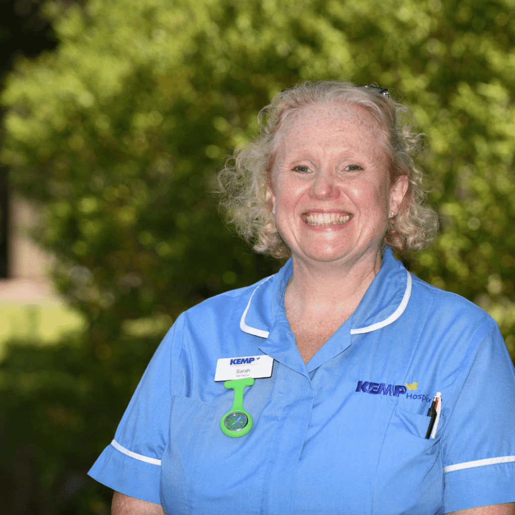 a nurse wearing light blue scrubs stands smiling looking directly at the camera with green shrubbery blurred in the background on a sunny day.