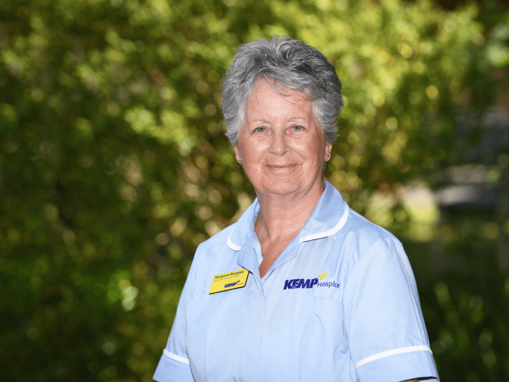 a nurse wearing light blue scrubs stands smiling looking directly at the camera with green shrubbery blurred in the background on a sunny day.