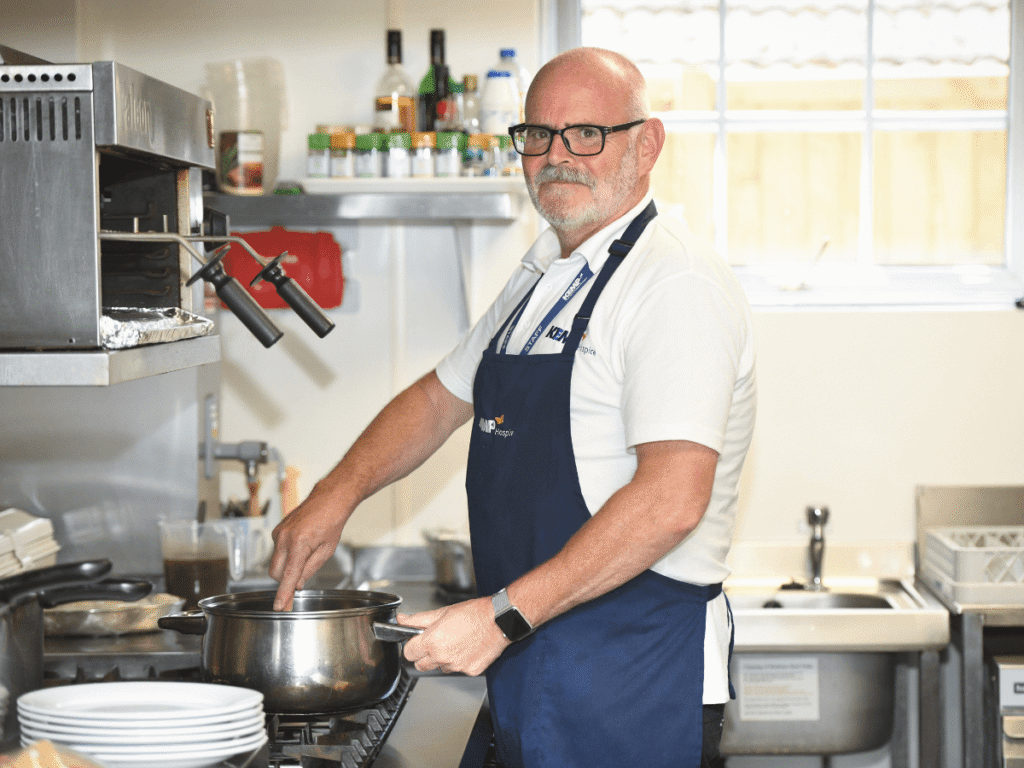 a gentleman chef stands next to a stove mixing food in a saucepan wearing a navy blue apron looking at the camera.