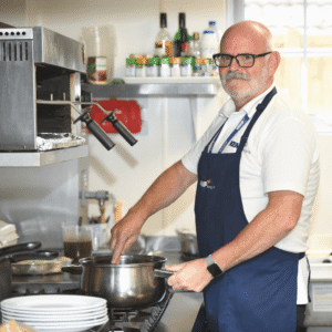 a gentleman chef stands next to a stove mixing food in a saucepan wearing a navy blue apron looking at the camera.