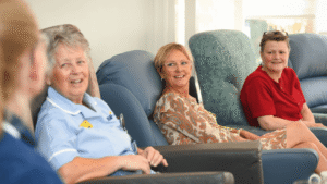 a nurse wearing light blue scrubs sits on a chair next to two ladies, each in charis. One lady wears a light brown dress and another wears a bright red top