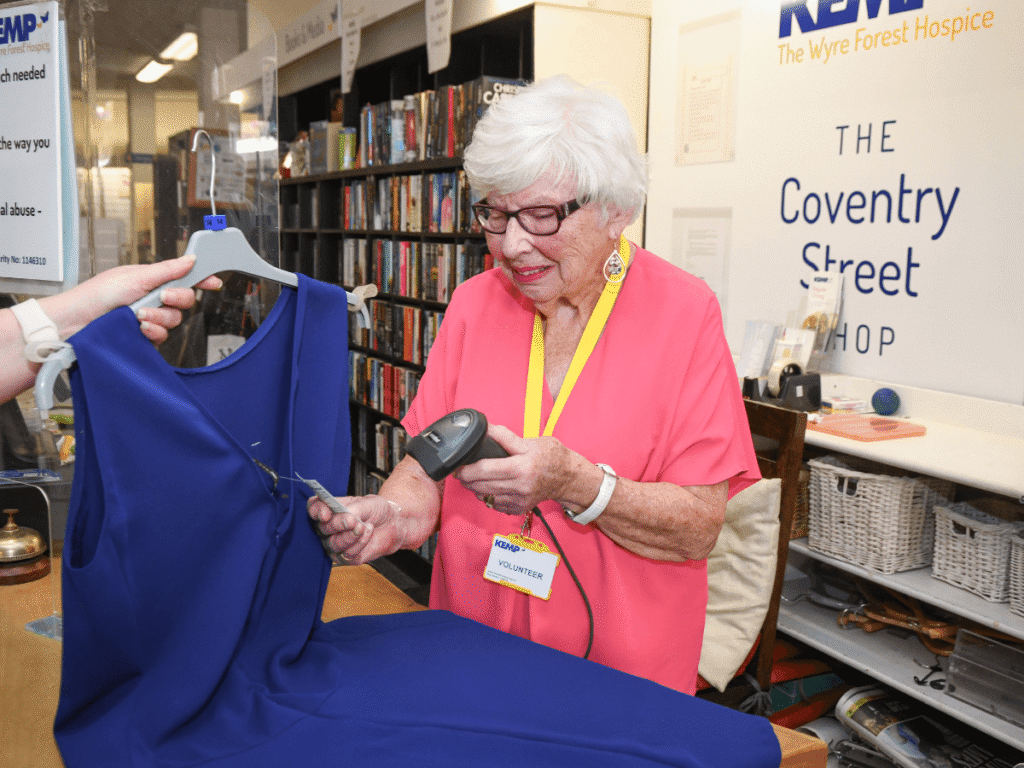 a lady in a pink top holds a barcode scanner whilst scanning the tag on a blue dress. She is stood by a till station with books and dvds behind her.