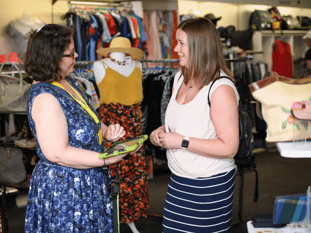 a lady in a pink top with a black backpack holds a blue dress she's taken from a shelf next to other clothes on a rail.