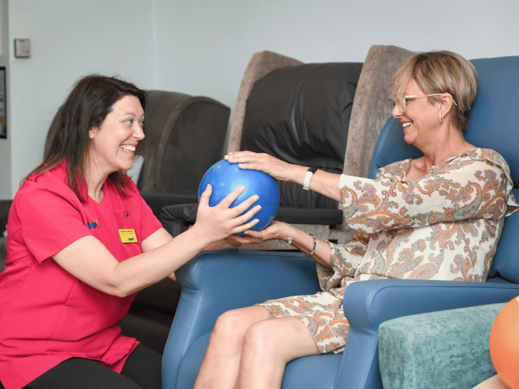 a lady in pink scrub top knelt down looking at a lady in an orange and cream top which holding a blue exercise ball