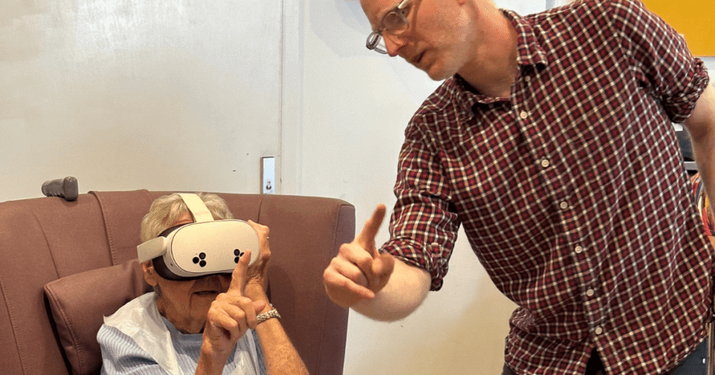 a lady sits in a chair wearing a virtual reality headset while a man stands next to her talking to her.